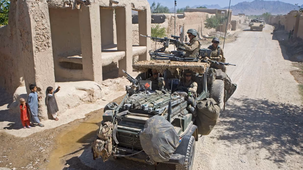 Alt-tekst: Een militair zit op een verhoging van zand en geeft een snoepje aan een meisje. Achter haar staat een jongen. 