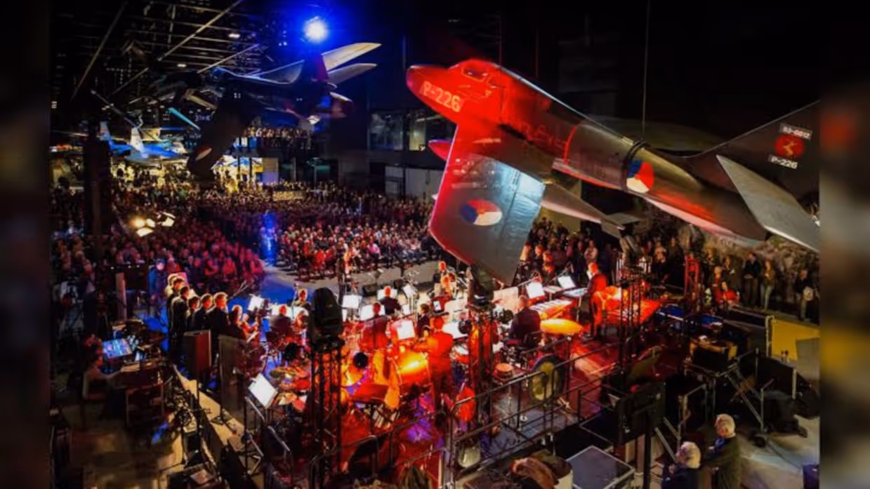 Overzicht van het podium met muzikanten en het publiek in het Nationaal Militair Museum. 