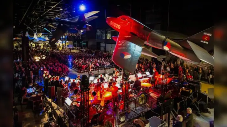 Overzicht van het podium met muzikanten en het publiek in het Nationaal Militair Museum. 