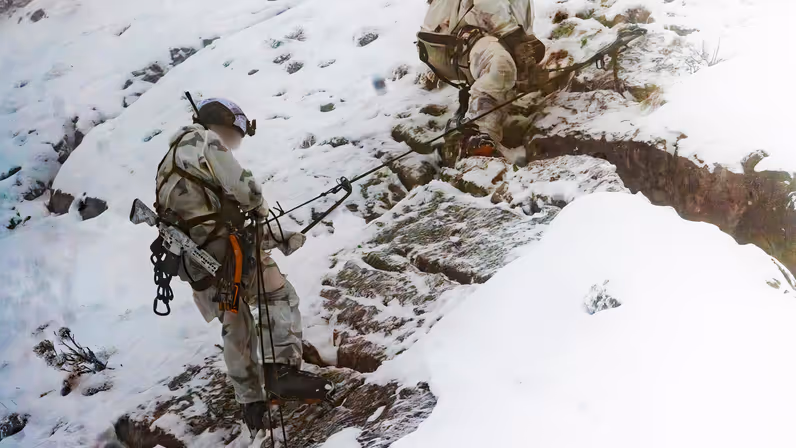 Twee militairen op een bergwand. Zij zijn gezekerd. De bovenste zit op een knie, de persoon daarachter daalt af met zijn wapen op de rug.