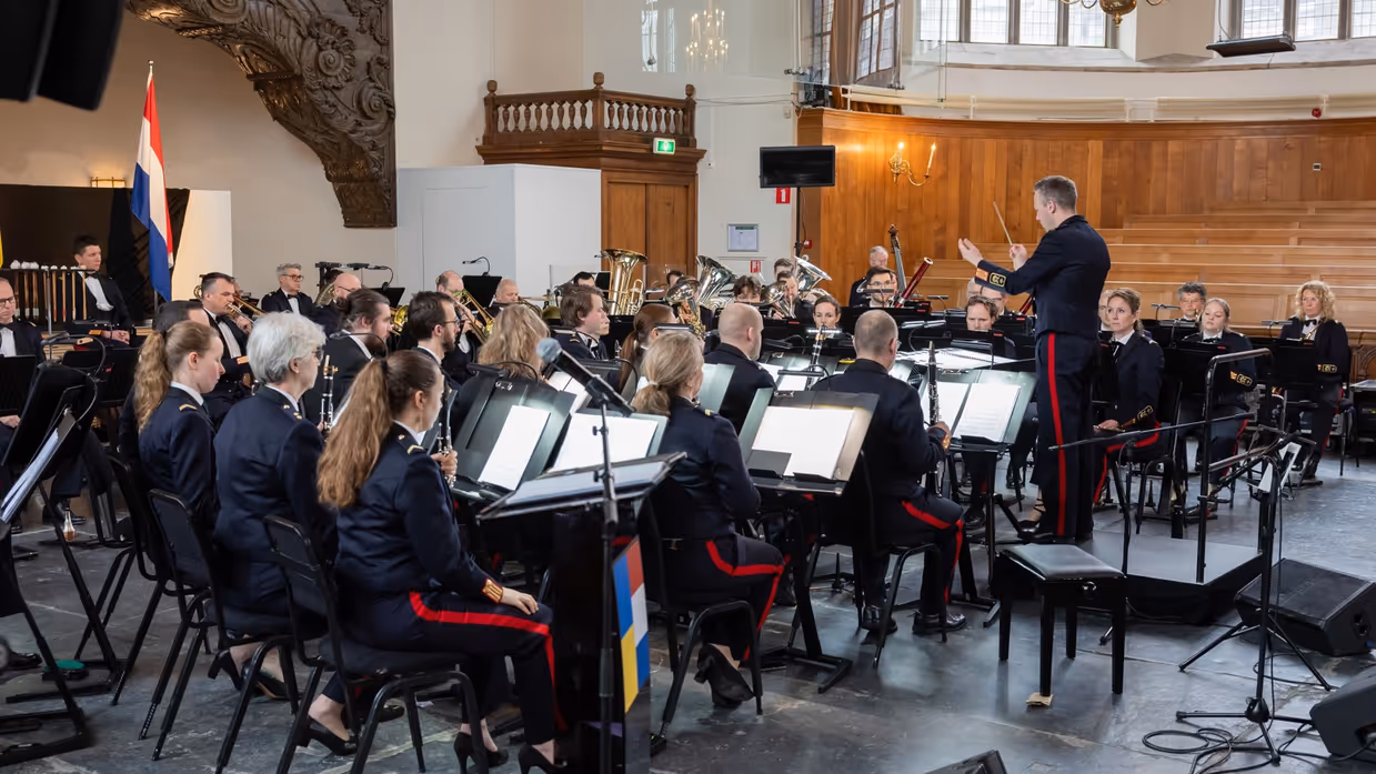 Dirigent staat voor militair orkest dat zittend speelt.