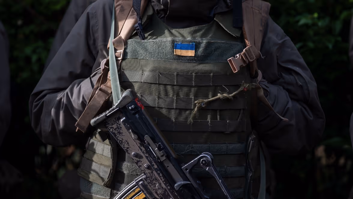 Close-up van de torso van een militair. Hij draagt een scherfwerend vest, een geweer en een Oekraiëns vlaggetje op zijn uniform.