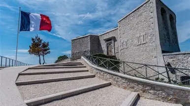 Een stenen monument met trappen, omringd door een hek, met een wapperende Franse vlag tegen een heldere hemel.
