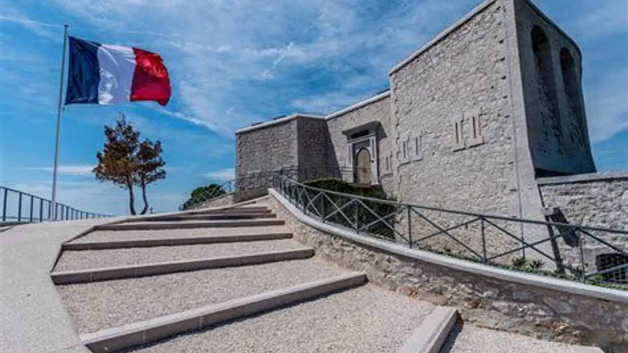 Een stenen monument met trappen, omringd door een hek, met een wapperende Franse vlag tegen een heldere hemel.