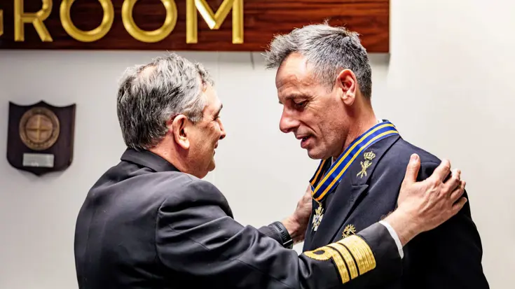 Viceadmiraal Jan Willem Hartman krijgt een onderscheiding om zijn nek gehangen van een man in pak, die zijn twee handen op de schouders van de militair heeft.