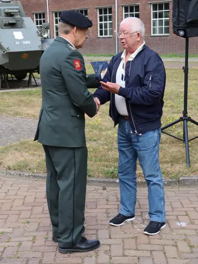 Een man in militair uniform reikt een medaille aan een oudere man in casual kleding, buiten bij een pantservoertuig.