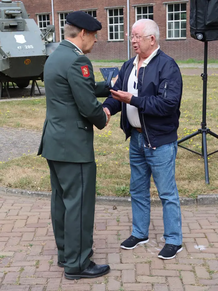 Een man in militair uniform reikt een medaille aan een oudere man in casual kleding, buiten bij een pantservoertuig.