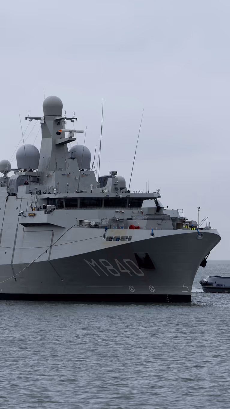 Een grijs marineschip vaart op een kalme zee, daarnaast een sleepboot. 