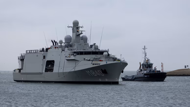 Een grijs marineschip vaart op een kalme zee, daarnaast een sleepboot. 