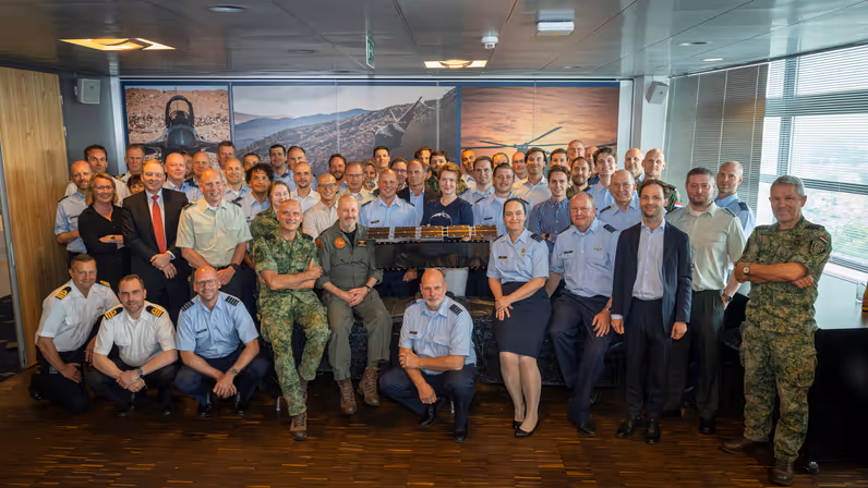 Groepsfoto van militairen en burgers rondom een schaalmodel van een satelliet.