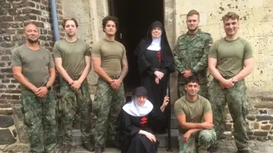 Zeven mannen in militaire kleding poseren met twee nonnen in zwarte gewaden voor een oude stenen deur op een kinderhoofdjesweg.
