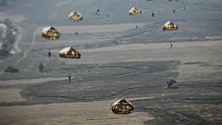 Meerdere parachutisten dalen met ronde parachutes af boven een uitgestrekt, landelijk landschap.