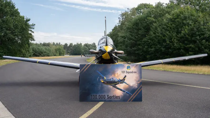 Een PC-7 op een taxibaan met daarvoor een bord met daarop het vliegtuig afgebeeld en de teksten ‘131 Squadron’ en ‘100.000 sorties’.