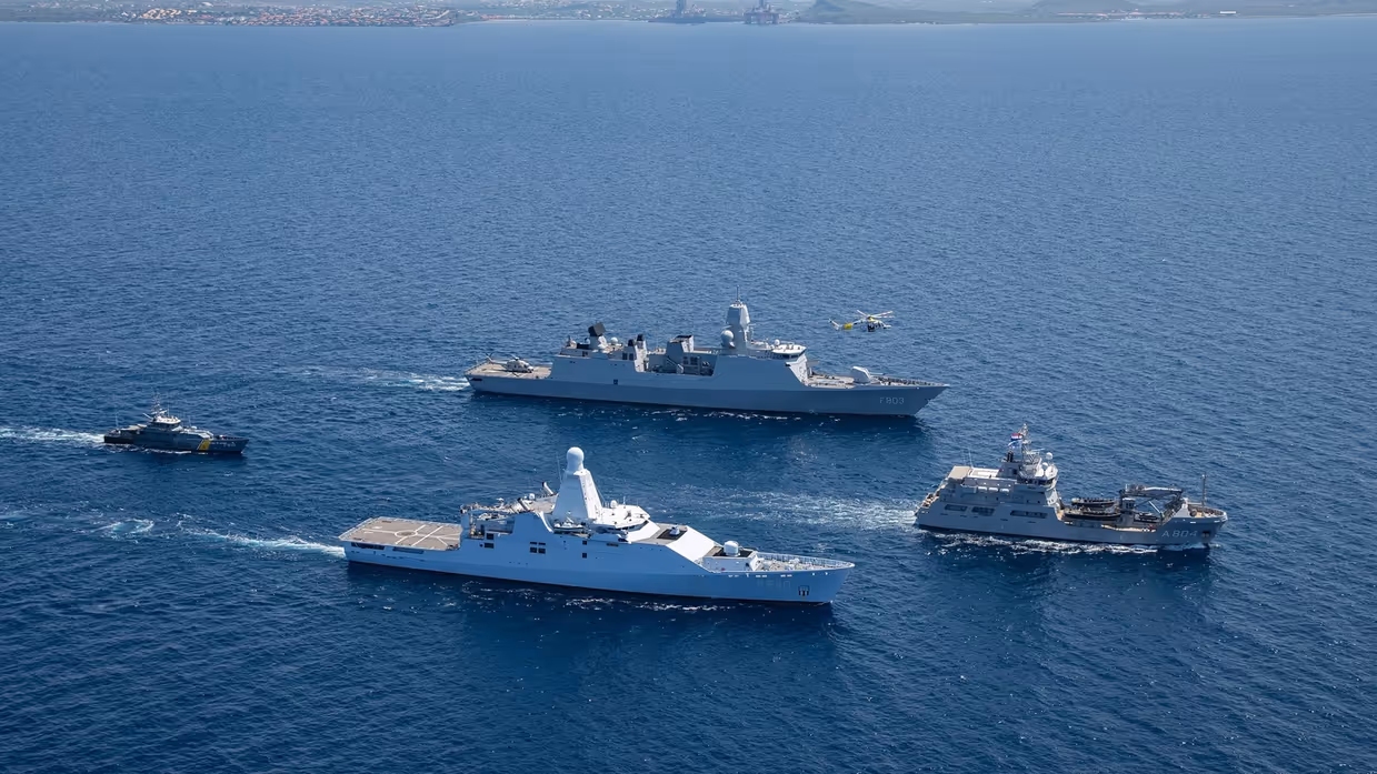 4 marinevaartuigen varen in formatie op open zee, met een helikopter erboven en land op de achtergrond.