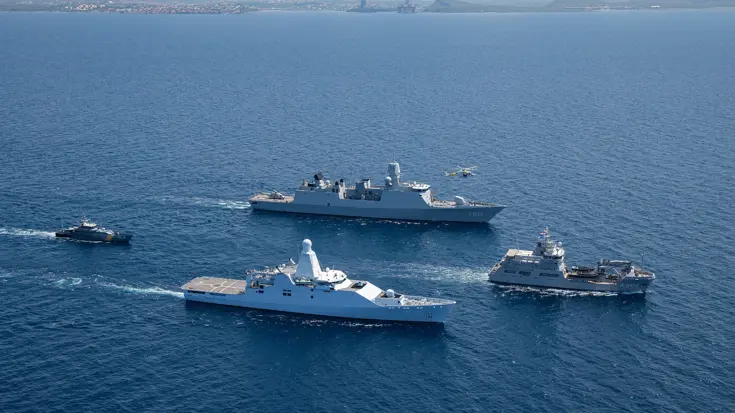 4 marinevaartuigen varen in formatie op open zee, met een helikopter erboven en land op de achtergrond.