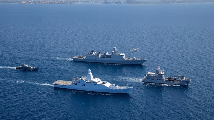 4 marinevaartuigen varen in formatie op open zee, met een helikopter erboven en land op de achtergrond.