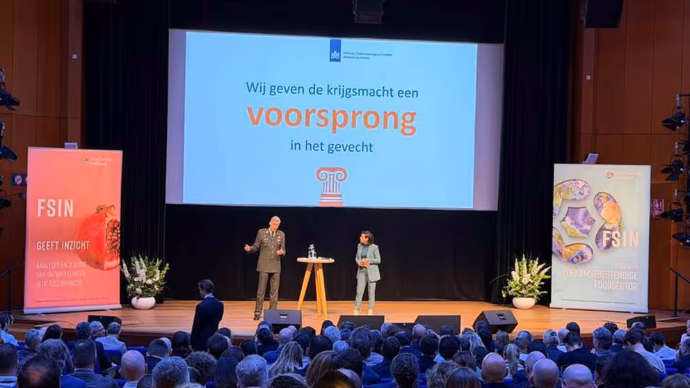 Luitenant-generaal Jan-Willem Maas staat op een podium met achter hem een groot scherm. Op de voorgrond van de foto achterhoofden van mensen in de zaal.