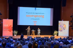 Luitenant-generaal Jan-Willem Maas staat op een podium met achter hem een groot scherm. Op de voorgrond van de foto achterhoofden van mensen in de zaal.