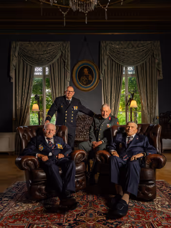 Vier veteranen zitten op donkere Chesterfieldbanken in de commandantenkamer op Bronbeek. De oude mannen kijken allemaal in de camera en dragen een net militair uniform in blauw en groen. 