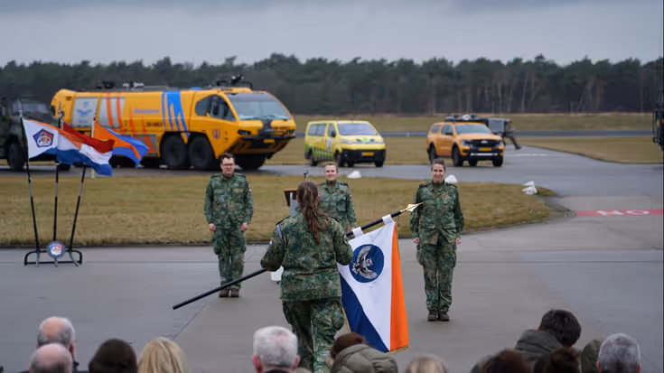 Vrouwelijke militair met vlag in de hand tegenover drie militairen op een platform. Achter hen militaire voertuigen waaronder een bluswagen. Op de voorgrond de achterhoofden van publiek.