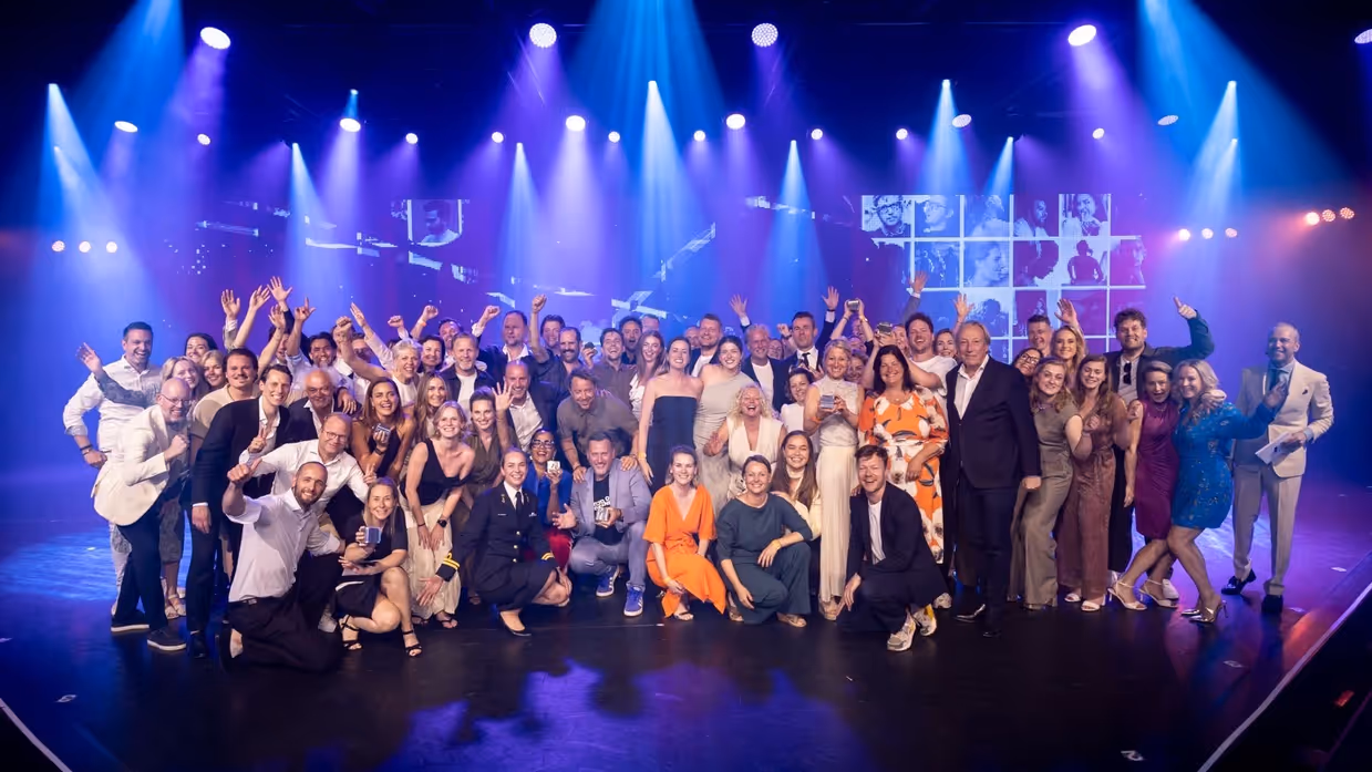 Een grote groep feestelijk geklede enthousiaste mensen op een blauw verlicht podium poseert voor de camera. Te midden van hen één vrouwelijke marineofficier.