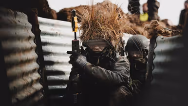Links: Luchtfoto van loopgraven op militair oefenterrein. Rechts: Militair met wapen hurkt in loopgraaf waarvan de wanden met golfplaten zijn bekleed. Op zijn helm een bos stro als camouflage.