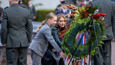 Yeşilgöz buigt samen met een jong meisje voorover naar een krans. Naast de krans staan twee landmachtmilitairen in dagelijks tenue.