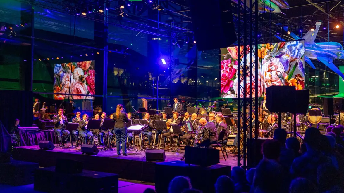 Een orkest speelt muziek in een donkere hal. Veteraan krijgt een ereteken uitgereikt.
