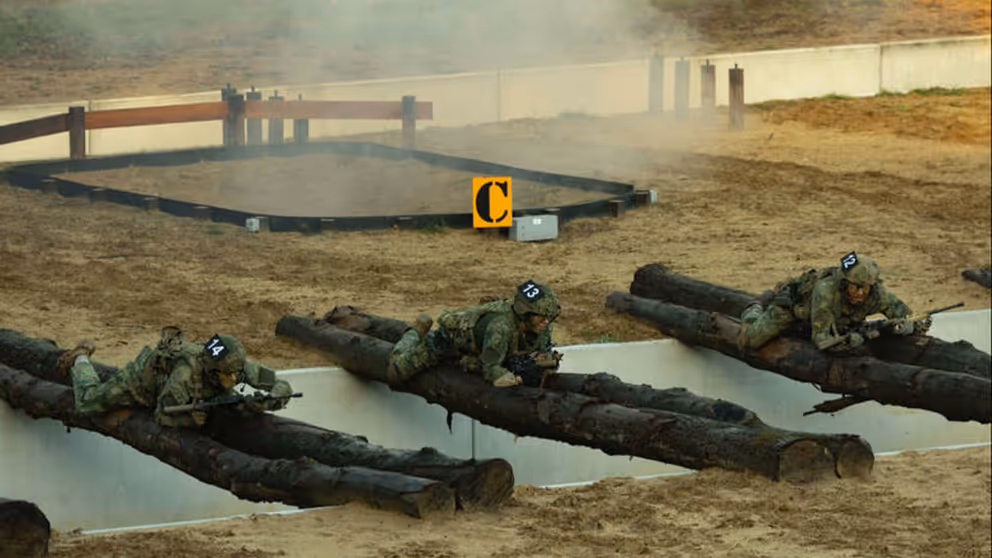 Een greppel met boomstammen die eroverheen liggen waar militairen overheen kruipen en rook oop de achtergrond.