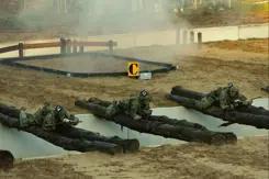 Een greppel met boomstammen die eroverheen liggen waar militairen overheen kruipen en rook oop  de achtergrond.