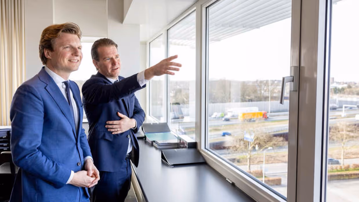 Twee mannen in pak kijken uit het raam over fabrieksterrein. Een van hen wijst iets aan.
