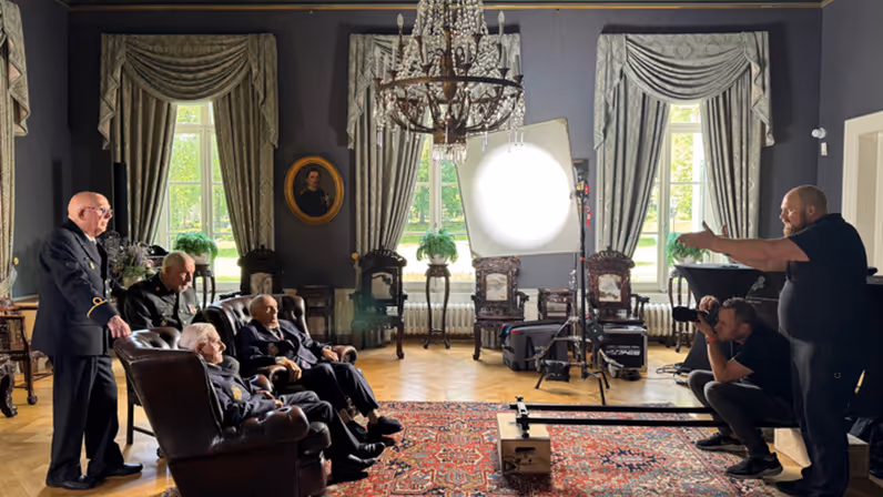 Regisseur Joas Burggraaf staat tegenover een groep oude veteranen. Zij staan in beeld van een camera onder een grote lamp in de commandantenkamer van Bronbeek.&nbsp;