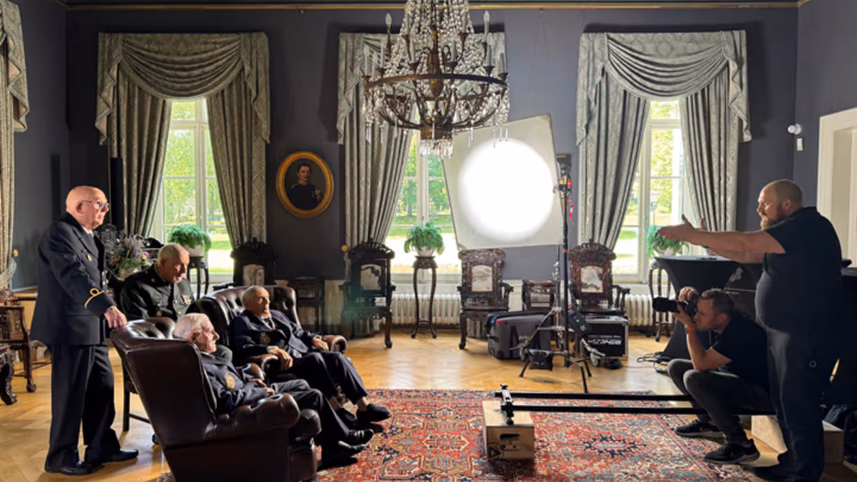 Regisseur Joas Burggraaf staat tegenover een groep oude veteranen. Zij staan in beeld van een camera onder een grote lamp in de commandantenkamer van Bronbeek.&nbsp;