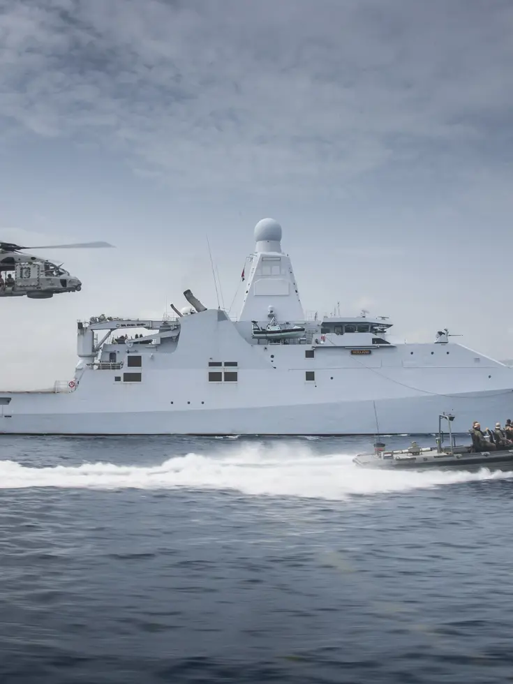 Schip op zee met boot die er naast vaart en een helikopter volgt.