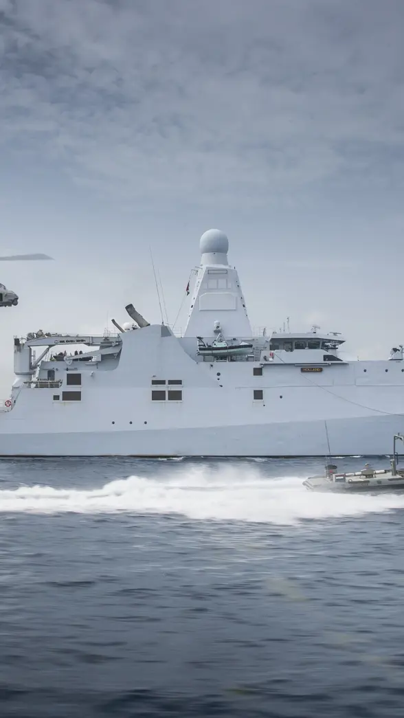 Schip op zee met boot die er naast vaart en een helikopter volgt.