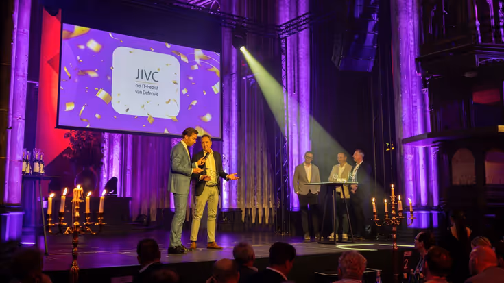 Twee mannen in pak op een podium, van wie een een microfoon vasthoudt. Ze staan in een statige, paars verlichte zaal.