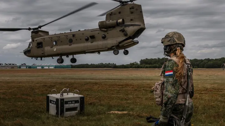 Prinses Amalia kijkt op een grasveld naar een vlak boven de grond vliegende Chinook. Ze draagt een gevechtstenue met helm en draagt in haar handen een bundel veiligheidsriemen.