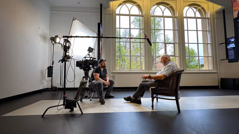 Mijnheer Boes zit op een stoel onder een microfoon. Tegenover hem zit regisseur Joas Burggraaf, die hem interviewt. 