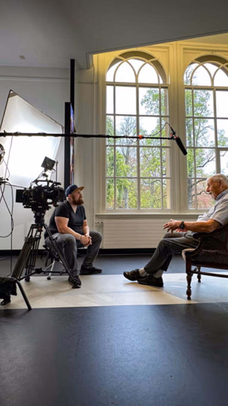 Mijnheer Boes zit op een stoel onder een microfoon. Tegenover hem zit regisseur Joas Burggraaf, die hem interviewt. 