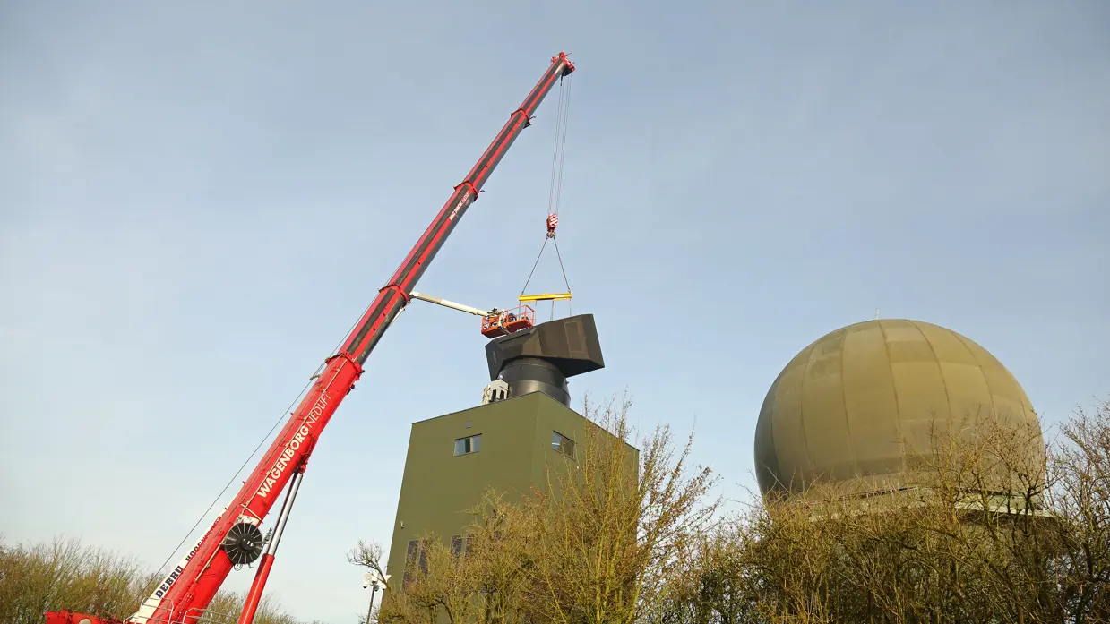 Links plaatst hijskraan een radar op een hoge toren. Rechts steekt een radarkoepel boven begroeiing uit.