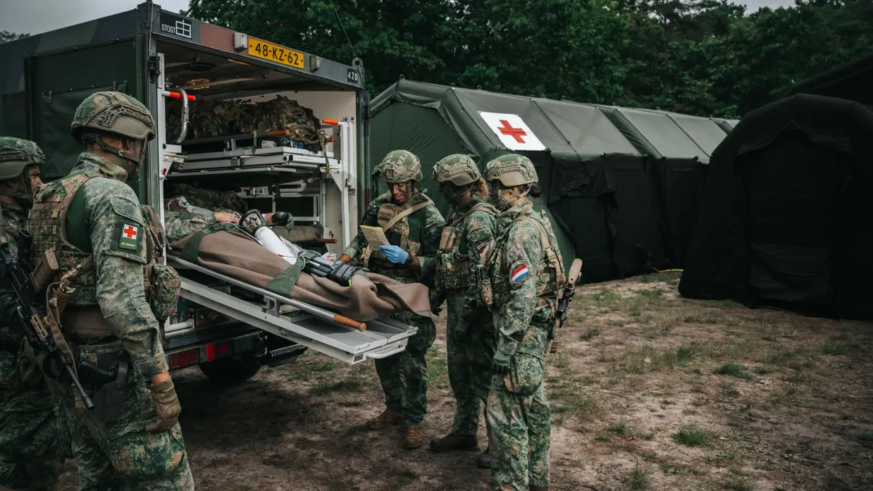 Alttekst: 5 militairen buigen zich over een gewonde collega die wordt ingeladen in een militair voertuig. 