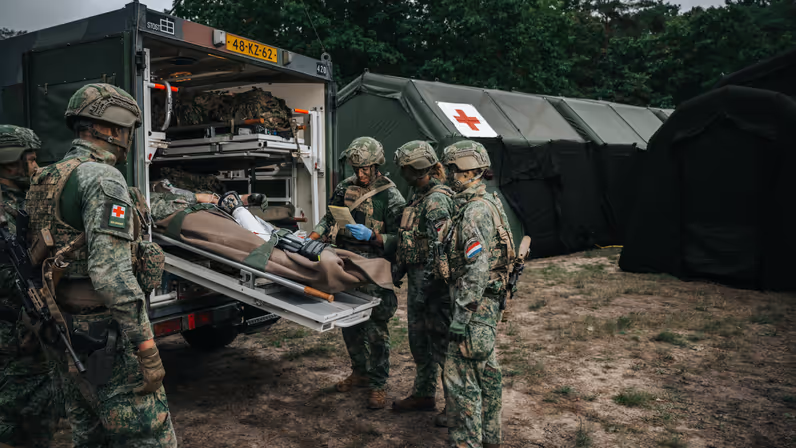 Alttekst: 5 militairen buigen zich over een gewonde collega die wordt ingeladen in een militair voertuig. 