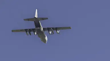 Een grijs militair transportvliegtuig met vier propellers vliegt tegen een heldere blauwe hemel met zichtbare motoren en staart.