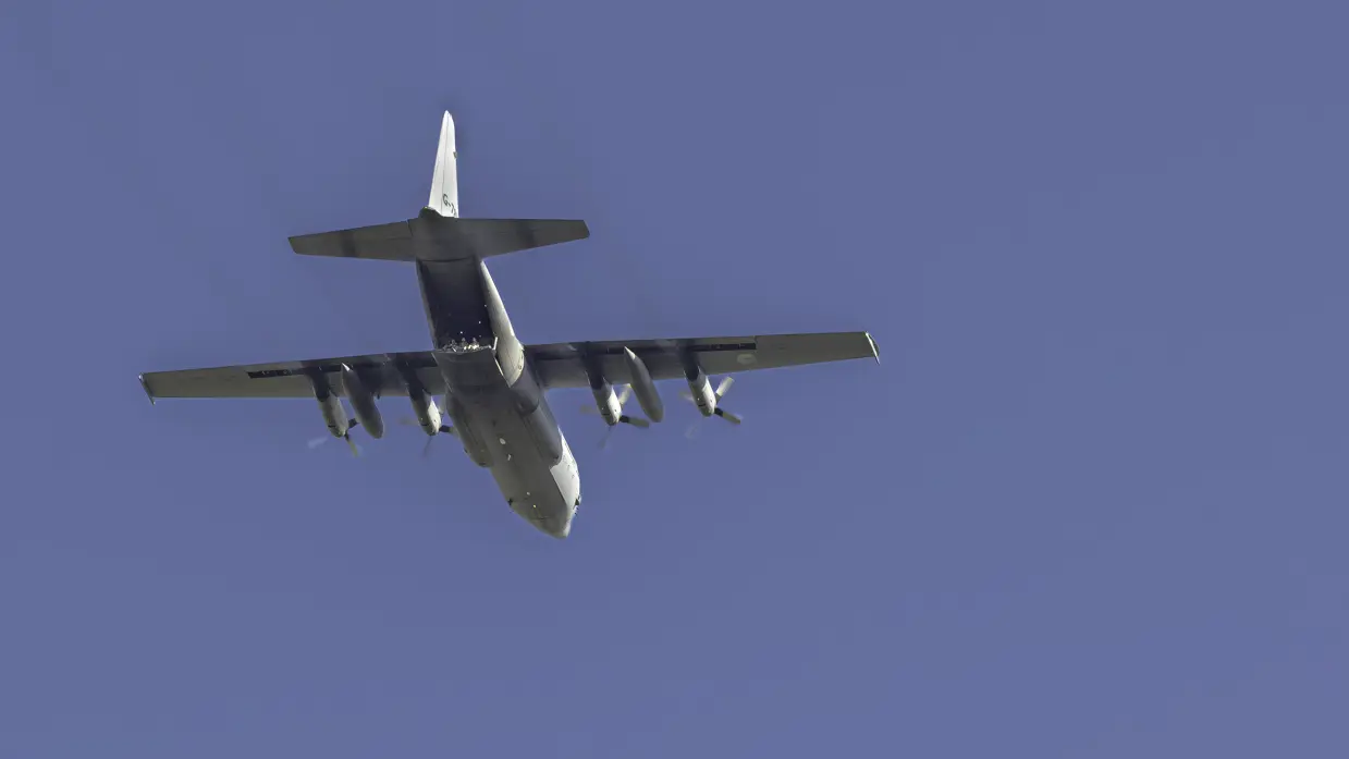 Een grijs militair transportvliegtuig met vier propellers vliegt tegen een heldere blauwe hemel met zichtbare motoren en staart.