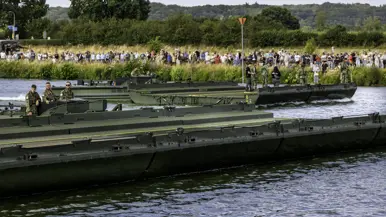 Drie groene militaire landingsvaartuigen varen over een rivier, met soldaten aan boord en een menigte toeschouwers op de oever. 