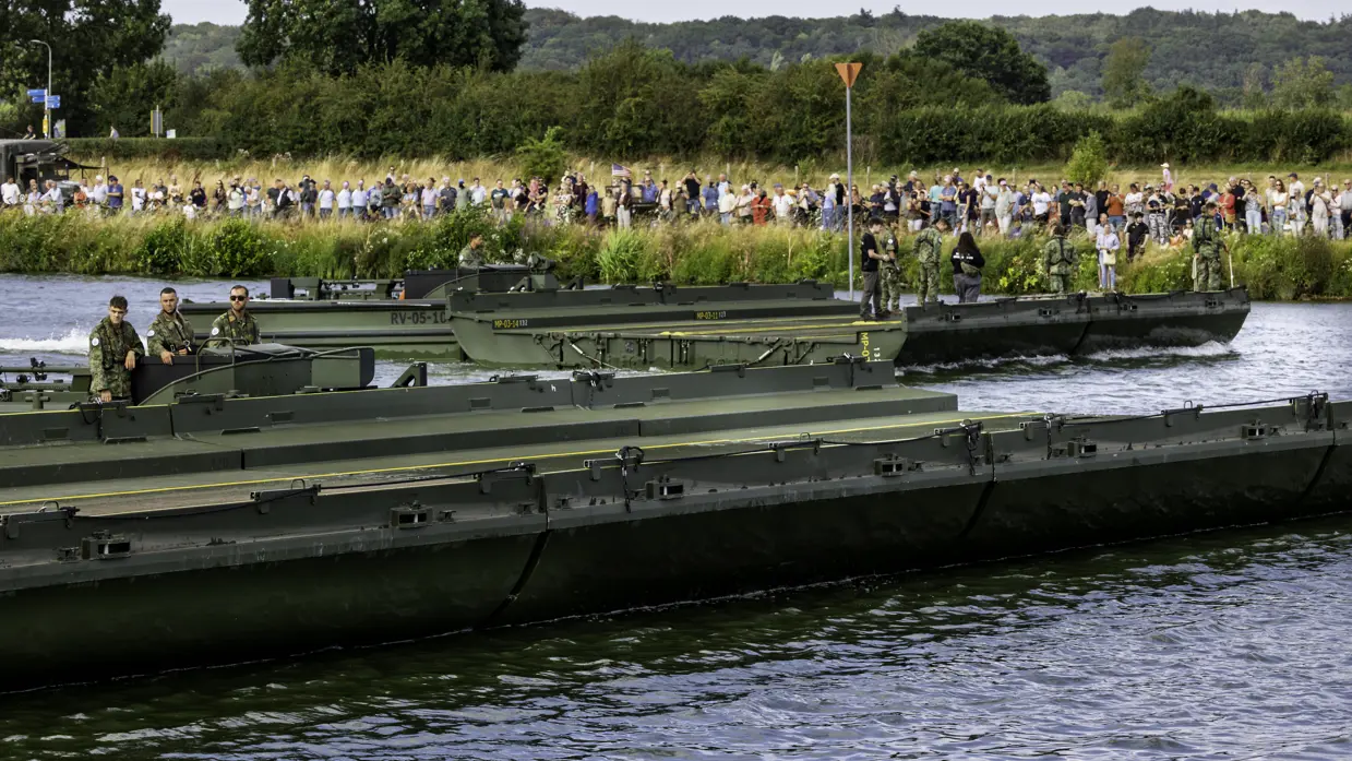 Drie groene militaire landingsvaartuigen varen over een rivier, met soldaten aan boord en een menigte toeschouwers op de oever. 