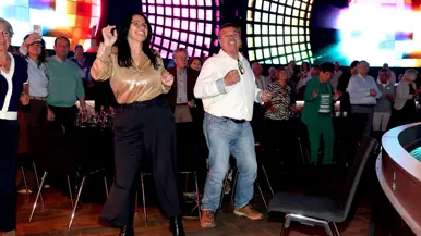Dansende mensen naast hun tafels en stoelen voor een disco-verlichte wand. 