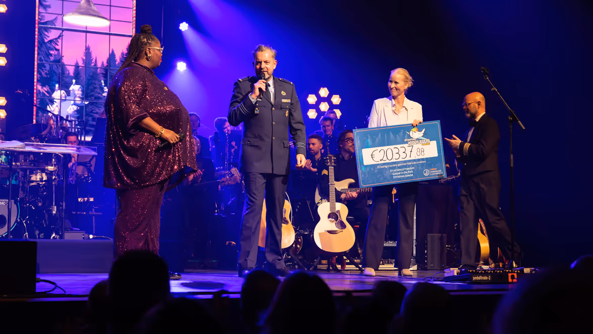 Op het podium staan zangeres Shirma Rouse, majoor Toine Jongbloets pratend door een microfoon en commodore Ellen Meeuwsen-Scholten met in haar handen een grote cheque. Op de achtergrond zit het orkest.