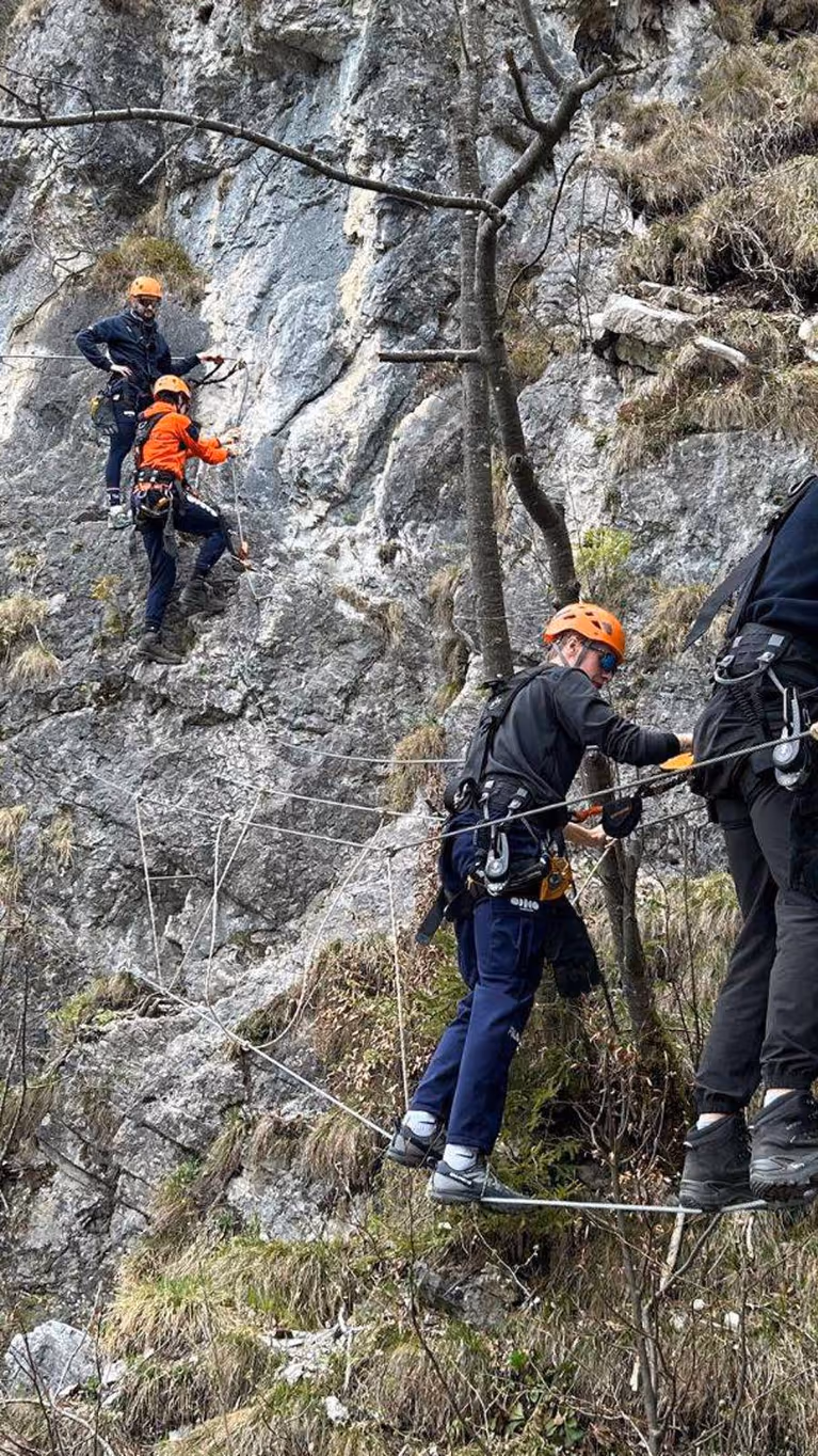 Drie mensen klimmen tegen een rotswand, beveiligd met touwen en helm. Ze dragen klimuitrusting, omringd door gras, bomen, en steile rotsen. Teamwerk en avontuur. 