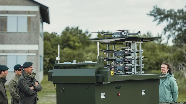 Drie militairen en een burger staan rondom een donkergroene, gelaagde kast. Daarin staat meerdere drone.